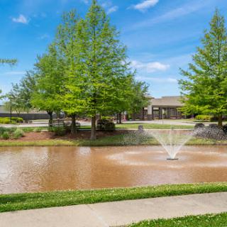 A serene park scene featuring a pond with a water fountain surrounded by lush greenery and clear blue skies.
