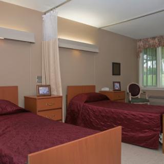 A cozy and well-lit bedroom featuring twin beds with burgundy covers, accented by wooden furniture and a view outside.