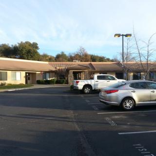 A small parking lot with parked cars in front of a single-story building, surrounded by trees during the daytime.