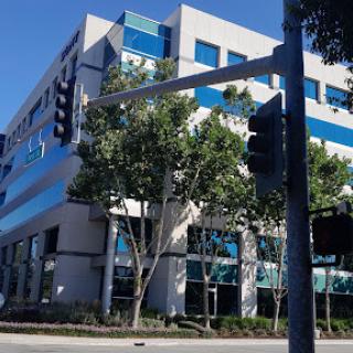 A modern office building featuring blue accents, surrounded by trees and a traffic signal.