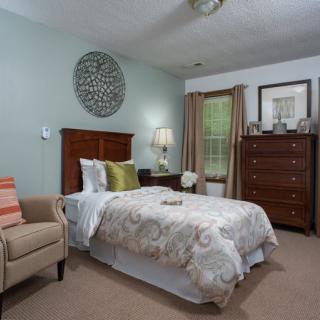 A cozy bedroom interior featuring a bed with patterned bedding, a wooden dresser, an armchair, and decorative pieces on the walls.