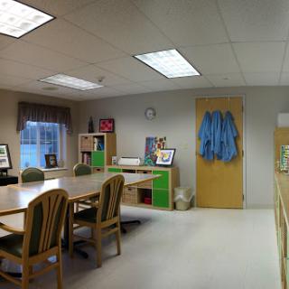 A well-lit office space featuring a meeting table surrounded by chairs, a desk area with shelves, and a door leading to another room.