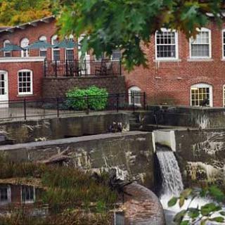 A historic brick building with a small waterfall flowing into a canal, surrounded by autumn foliage.