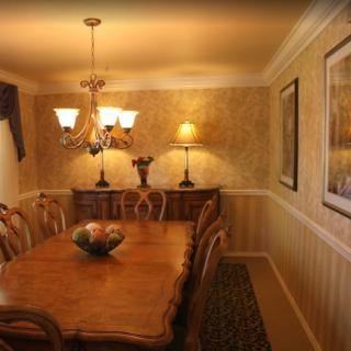 A warmly lit dining room with a wooden table and elegant chairs, featuring a chandelier and wall art.