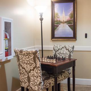 A cozy corner with a chess table and patterned chairs, softly lit by a nearby lamp, featuring a decorative painting on the wall.