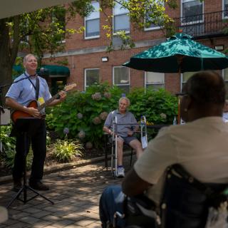 A guitarist performs for an audience of seniors enjoying a sunny day outdoors.