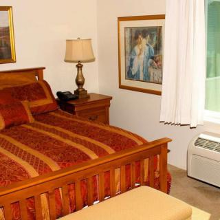 A cozy bedroom featuring a wooden bed with patterned bedding, a bedside lamp, framed artwork on the wall, and white curtains covering the window.