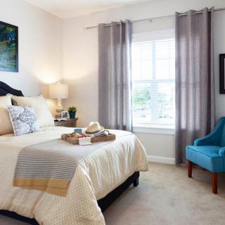A cozy bedroom with a neatly made bed, a blue chair, and soft lighting from a lamp by the window.