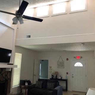 A modern living room with high ceilings and a ceiling fan, featuring a fireplace, television, and sofa. Natural light filters in through multiple small windows above.