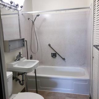 A compact bathroom featuring a bathtub with a showerhead, a small sink, and a toilet. There is a mirror above the sink and a grab bar in the bathtub area for safety.