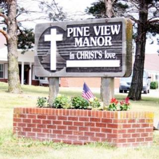 A sign for Pine View Manor featuring a cross, surrounded by a brick base and an American flag.