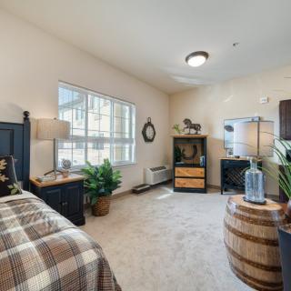 A cozy bedroom with natural light, featuring a bed, window, decorative plants, and wooden furniture.