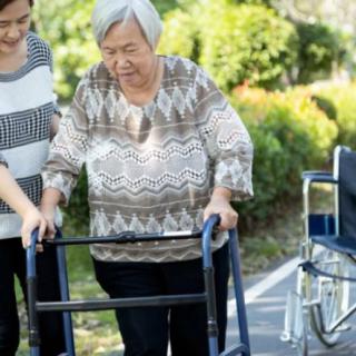A caregiver helps an elderly woman using a walker in an outdoor setting.