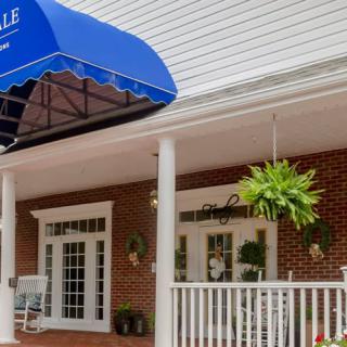 A building entrance featuring a blue awning, porch with columns, hanging plants, and white railings.