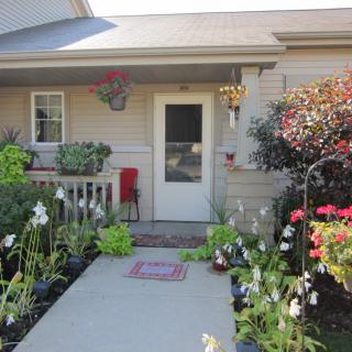 A charming house entrance adorned with vibrant flowers and lush greenery along a concrete pathway.