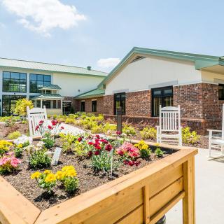 A sunny outdoor view of a building with a landscaped garden, featuring colorful flowers and white rocking chairs.