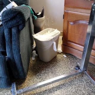 A cluttered corner showing a trash can next to some furniture and a wooden cabinet.