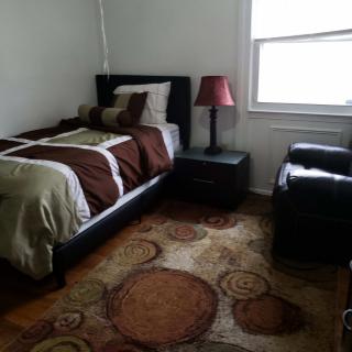 A cozy bedroom with a single bed, armchair, and nightstand, featuring a colorful rug and a lamp by the window.