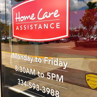 A window sign for Home Care Assistance showing business hours and contact information.