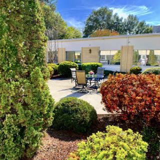 A serene outdoor garden setting featuring neatly trimmed bushes, a cozy patio area with furniture, and a clear blue sky.