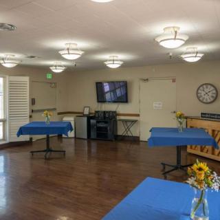 A well-lit room with tables set with blue tablecloths, a wall-mounted television, flowers in a vase, and a clock on the wall.