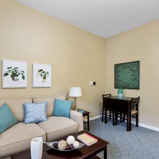 A cozy living room with a beige sofa, decorative pillows, and a coffee table, accented by wall art and a small dining area.