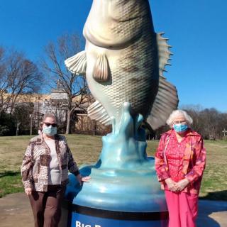 Two people wearing masks stand beside a large fish statue outdoors on a sunny day.