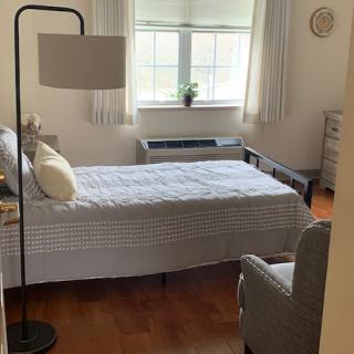 A minimalist bedroom with a single bed, a lamp, and an armchair next to a window.