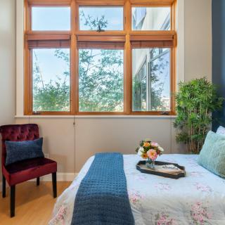 A cozy bedroom with a large window, featuring a bed adorned with floral bedding, a side chair, and vibrant decorative elements.