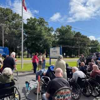 A gathering of elderly individuals in wheelchairs attending an outdoor event, with speakers addressing the crowd near a flag.
