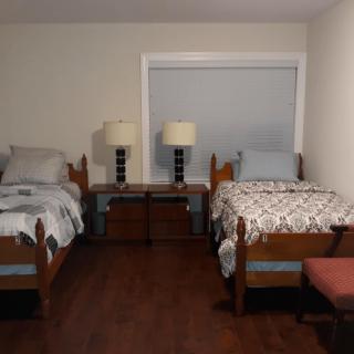 A cozy bedroom with two twin beds, each adorned with patterned bedding and separated by a nightstand with two lamps. The room features a large window and hardwood flooring.