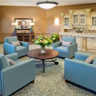 A cozy living room setting featuring blue armchairs arranged around a round wooden table with a floral arrangement, set on a patterned carpet.