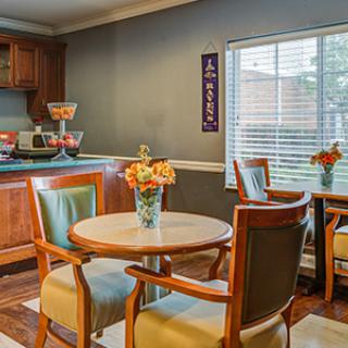 A cozy dining area with wooden furniture, decorated with vases of flowers, and illuminated by natural light from a window.
