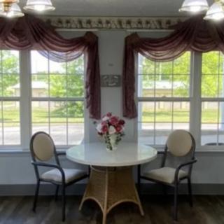 A cozy dining area with a round table and two chairs by large windows adorned with elegant curtains, featuring a floral centerpiece.