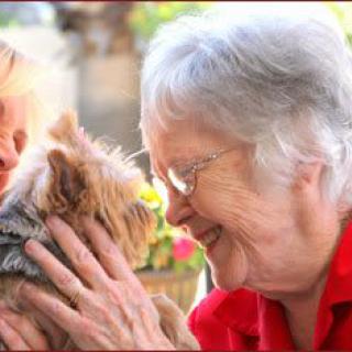 An elderly woman smiling and showing affection to a small dog outdoors.