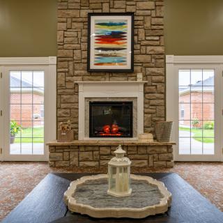 A cozy living room with a central stone fireplace, flanked by two large windows and decorated with colorful artwork.