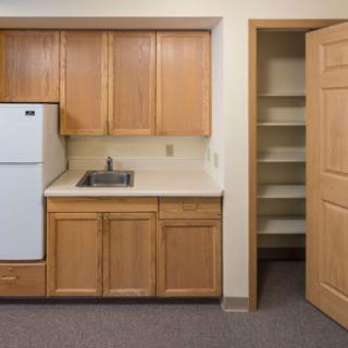 A small kitchen with wooden cabinets, a refrigerator, and an open pantry door.