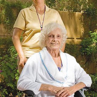 A caregiver stands behind an elderly woman seated in a wheelchair, both sharing a moment outdoors.