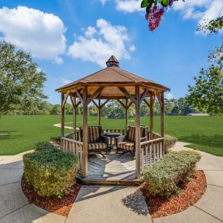 A wooden gazebo surrounded by lush greenery and a paved walkway on a sunny day.