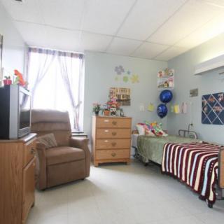 A cozy hospital room with a single bed, armchair, dresser, and television.