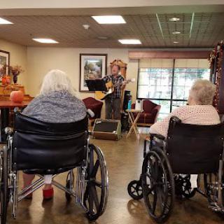 A musician performs for elderly residents in a nursing home, with several audience members seated in wheelchairs.