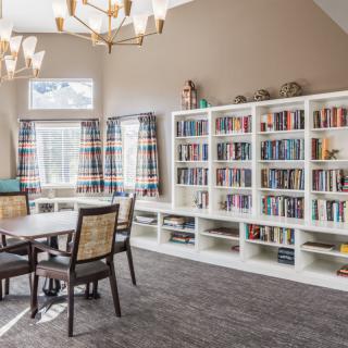 A cozy library room with white bookshelves filled with books, a table and chairs, and large windows with plaid curtains.