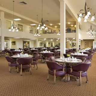 A spacious dining room with multiple round tables and chairs, featuring chandeliers and a carpeted floor.