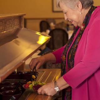 A woman in a pink jacket serves herself at a buffet table indoors.