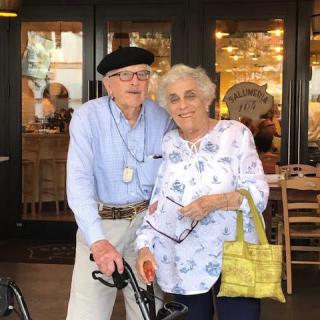 An elderly couple standing together in front of a store, one holding a cane.