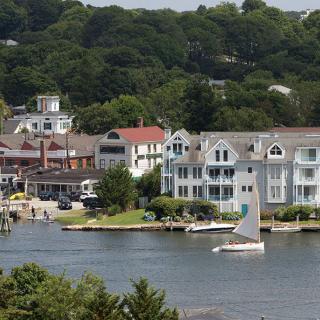 A charming coastal town featuring a sailboat gliding on the water, surrounded by picturesque buildings and lush greenery.