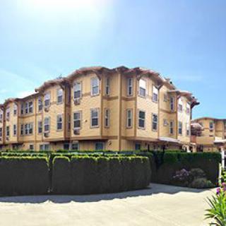 A sunny day view of a multi-story residential apartment building with a neatly trimmed hedge and fence.