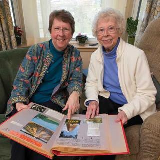 Two women sitting on a couch, smiling and looking through a scrapbook.