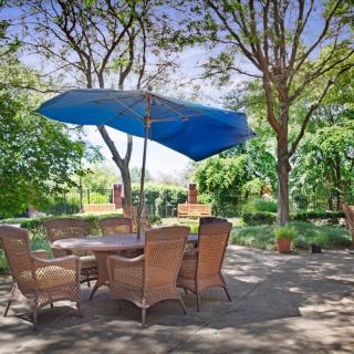 A peaceful outdoor patio with wicker chairs and a table under a blue umbrella, surrounded by lush greenery.