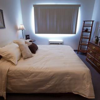 A cozy bedroom featuring a neatly made bed with white linens and soft pillows, illuminated by a bedside lamp near a shaded window.